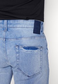 Los pantalones vaqueros de mezclilla azul claro presentan un diseño clásico de cinco bolsillos, una cinturilla visible y una etiqueta de cuero azul oscuro con la marca en la parte trasera.
