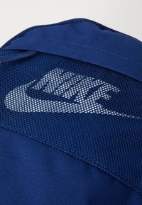 Nike kuprinė su mėlynu, tekstūruotu tinklu ir dideliu baltu logotipu. Medžiaga yra patvari su lygiomis detalėmis.