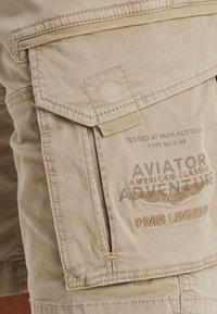 Beige cargo shorts with multiple pockets, textured fabric, reinforced stitching, and branding, including "Aviator Adventure" and "PME Legend."
