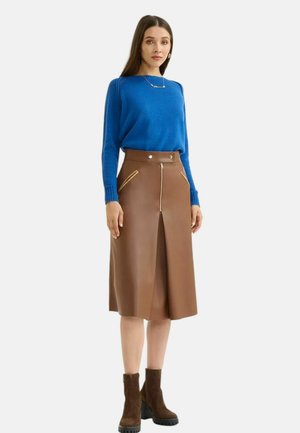 Gonna in ecopelle marrone con zip frontale, due tasche laterali e design plissettato; abbinata a un maglione blu e stivali alla caviglia marroni.