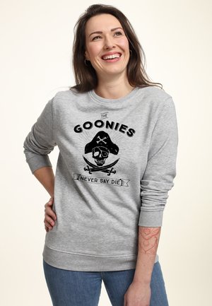 Smilende kvinde med mørkt hår iført en grå sweatshirt med piratskalle og korslagte knogler, teksten "The Goonies Never Say Die" og blå jeans.