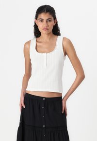 White sleeveless top with a textured pattern, scoop neckline, and decorative buttons. Matches with a black, button-up skirt.