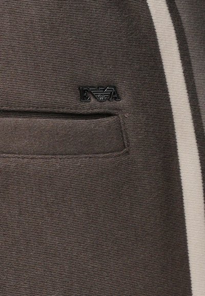 Κοντινή λήψη σκούρας καφέ υφάσματος με μια μικρή τσέπη και ένα μαύρο μεταλλικό λογότυπο που γράφει "EVA" πάνω από την τσέπη σε ένδυμα.