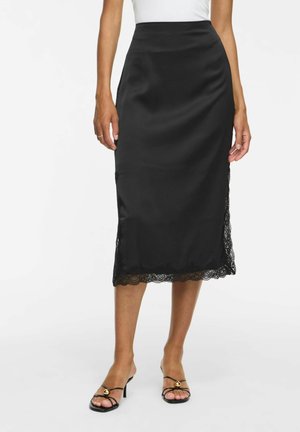 Femme portant une jupe midi en satin noire avec un bord en dentelle à l'ourlet, associée à des sandales noires à talons et à brides.