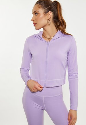 Frau mit braunem Pferdeschwanz, die einen hellvioletten Hoodie mit Reißverschluss und passende Leggings trägt und nach links vor einem schlichten Hintergrund schaut.