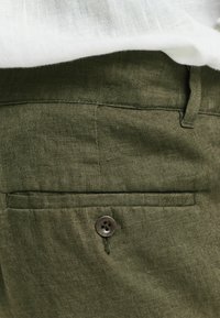 Olivgrüne Leinenhosen verfügen über eine Gesäßtasche mit einem einzelnen braunen Knopf und feiner Textur, mit einer sichtbaren Naht entlang der Taschenkante.