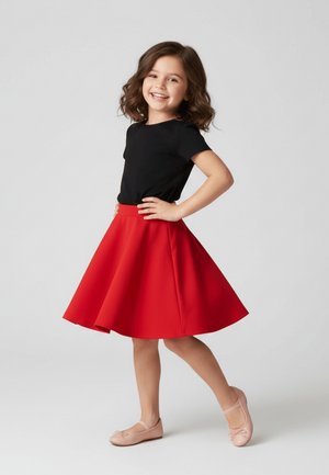 Młoda dziewczyna uśmiechająca się, stojąca z rękami na biodrach, ubrana w czarną koszulkę, czerwoną spódnicę i różowe baleriny na jasnym tle.