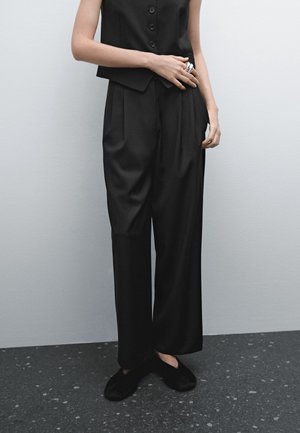 Personne portant un pantalon noir ample, un gilet noir sans manches boutonné, des chaussures noires duveteuses sans lacets, debout contre un mur gris clair.