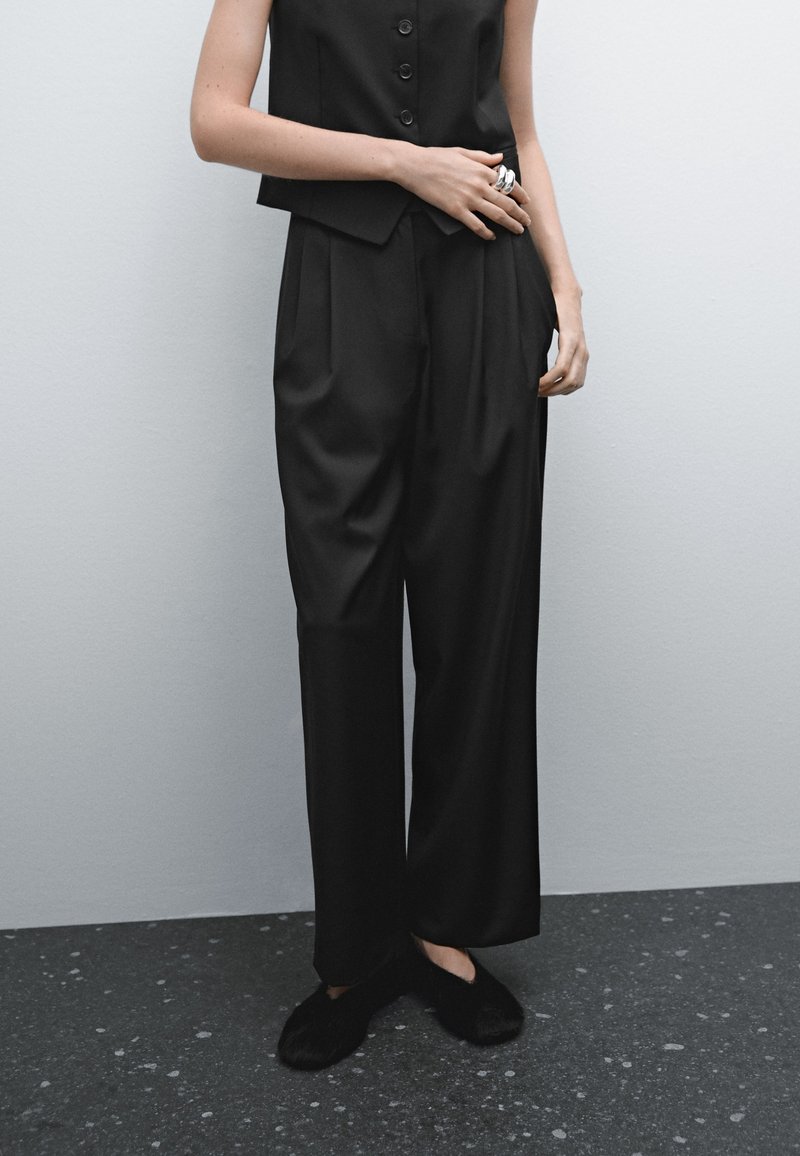 Personne portant un pantalon noir ample, un gilet noir sans manches boutonné, des chaussures noires duveteuses sans lacets, debout contre un mur gris clair.