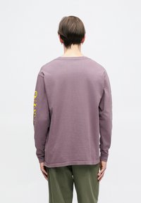 Ilgi rankoviai marškiniai iš neryškios levandų spalvos medvilnės su laisvu pritaikymu. Dešinėje rankovėje yra geltonas grafiškas spaudinis. Atrodo iš nugaros.