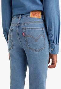 Jasnoniebieskie dżinsy z widocznymi tylnymi kieszeniami, pomarańczowymi szwami, czerwoną metką Levi's i skórzaną naszywką Levi Strauss & Co. na talii, noszone z dżinsową koszulą.