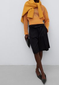 Oranger Strickpullover, kombiniert mit einem schwarzen, plissierten Rock, schwarzen spitz zulaufenden Schuhen und einer schwarzen Clutch. Sichtbare strukturierte Materialien.