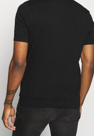 Mężczyzna w prostym czarnym krótkim rękawie i czarnych jeansach, ukazujący wytatuowane prawe przedramię, zwrócony tyłem do kamery.