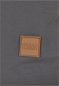 Tumehall kangas, millel on ruudukujuline heledat pruunikat nahast plaastriga, millel on reljeefne tekst "URBAN CLASSICS" ja õmmeldud servad. Lihtne ja kaasaegne disain.