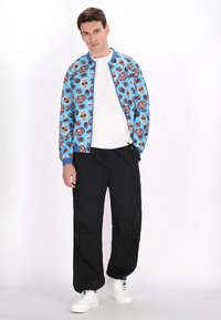 Jachetă bomber albastră cu un model floral și cu cranii pe toată suprafața, asortată cu pantaloni negri și adidași albi. Ansamblu simplu și casual.