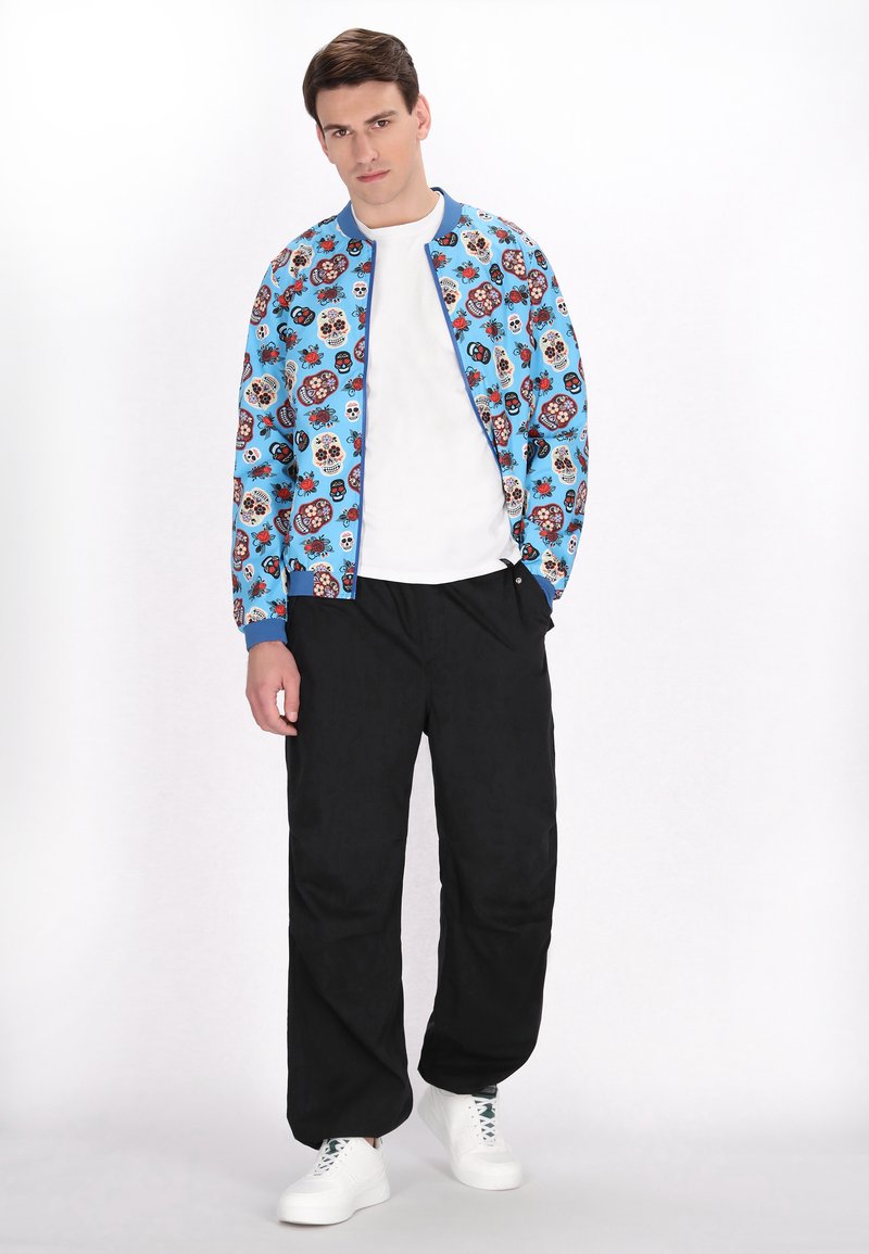 Jachetă bomber albastră cu un model floral și cu cranii pe toată suprafața, asortată cu pantaloni negri și adidași albi. Ansamblu simplu și casual.