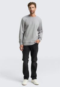 Felpa grigia realizzata in tessuto con finitura texture, abbinata a jeans scuri e sneakers bianche. Il modello si trova in piedi con le mani nelle tasche.