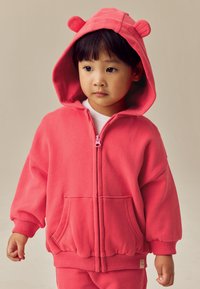 Lyserød hættetrøje med lynlås, bjørneører, lange ærmer, store forlommer og ribbet kant ved ærmerne. Blødt stof med en glat tekstur.