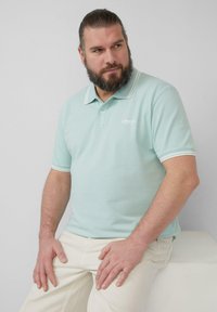Hellblaues Poloshirt mit weißen Kragen- und Ärmelstreifen, aus Baumwolle, mit kleinem Logo auf der Brust. Getragen mit cremefarbenen Hosen.