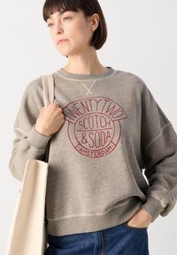Sivý bavlnený sweatshirta s okrúhlym marhuľovým logom s nápisom "TWENTY TWO SCOTCH & SODA AMSTERDAM." Má voľný strih a rebrované manžety.