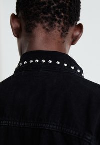 Black denim jacket with a studded collar, featuring metal studs along the seam and a textured fabric finish.