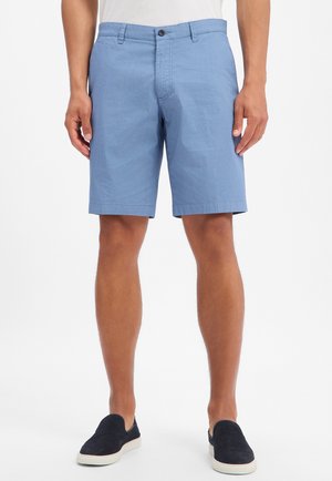 Mann trägt hellblaue, strukturierte Shorts, weißes Hemd und schwarze Slipper, steht vor weißem Hintergrund.