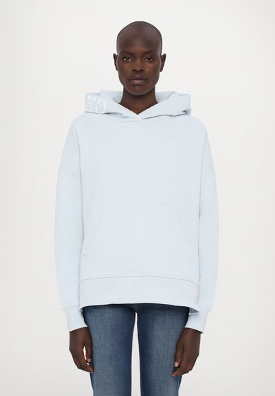 Haut à capuche bleu clair en tissu doux, avec une poche avant et un cordon de serrage au niveau de la capuche. L'épaule gauche présente un détail de logo blanc.