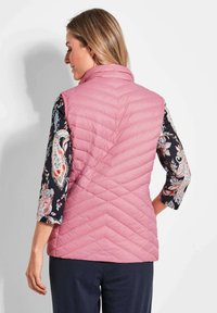 Rosa gesteppte Weste mit hohem Kragen, versehen mit einem Zickzackmuster. Getragen über einer dunklen Bluse mit Blumenmuster und Dreiviertelärmeln.