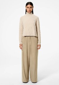Maglione lavorato a maglia beige con colletto alto e motivo a diamante, abbinato a pantaloni beige larghi. Texture liscia e design cropped.
