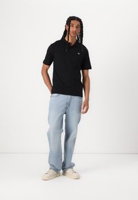 Svart kortermet polo skjorte med krage og lite brodert logo, kombinert med lyseblå, løstsittende jeans og hvite sneakers.