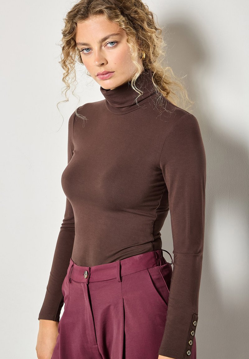 Brązowy sweter z golfem wykonany z miękkiego materiału, z długimi rękawami z guzikami przy mankietach, zestawiony z wysokim pasem śliwkowych spodni.