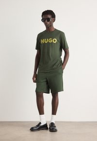 Grøn bomulds t-shirt med gul "HUGO" tekst, parret med matchende grønne shorts. Modellen bærer sorte solbriller og loafers med hvide sokker.