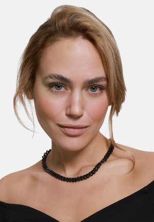Zwarte ketting hangt om de nek en heeft met elkaar verbonden, ronde schakels. Het model heeft een natuurlijke make-up en draagt een top zonder schouderbanden.