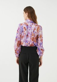 Leichte Bluse mit floralem Muster in lila, rosa und braunen Tönen; ausgestattet mit Puffärmeln und einem Kragen; kombiniert mit schwarzen, taillierten Hosen.