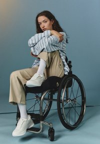 Baskets blanches, pantalons beige ajustés et une chemise oversize à rayures bleues. Modèle assis dans un fauteuil roulant noir sur fond bleu.