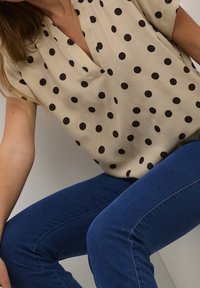 Haut beige à manches courtes avec des pois noirs, présentant un décolleté en V. Porté avec un jean en denim bleu, assis sur un fond neutre.