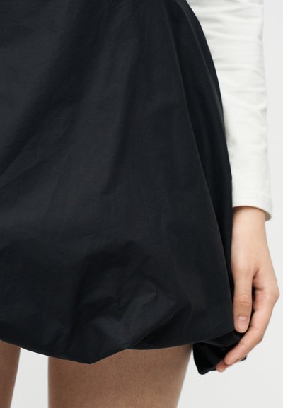 Jupe ballon noire portée sur des jambes nues, avec une main tenant délicatement l'ourlet ; haut blanc à manches longues partiellement visible.