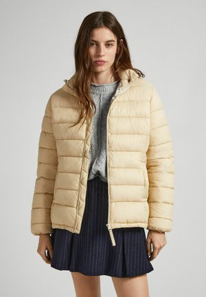 Giacca a vento beige con chiusura a zip e cappuccio, caratterizzata da una texture trapuntata e polsini a coste, indossata sopra un maglione grigio e una gonna a righe scure.
