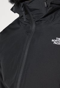 Veste noire zippée avec col montant et cordons de capuche, avec le logo blanc « The North Face » sur la poitrine.
