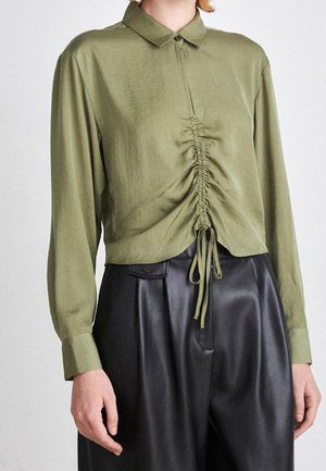 Persona que lleva una camisa de manga larga verde oliva con cordón fruncido en la parte delantera y pantalones plisados de talle alto en negro.