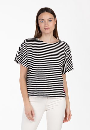 Vrouw met recht bruin haar, gekleed in een zwart-wit gestreept kort mouw shirt en witte broek, staand tegen een effen achtergrond.
