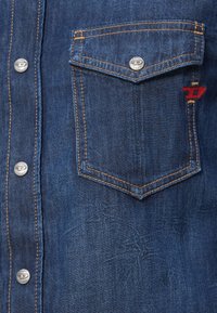 Tumši zila džinsa krekls ar kabatu, kas aizveras ar sudraba pogu, kontrastējošām dzeltenām šuvēm un nelielu sarkanu logotipu uz kabatas.