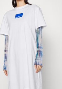 Weißes, übergroßes T-Shirt-Kleid mit blauem Grafiklogo, kombiniert mit durchsichtigen langen Ärmeln im Batikmuster in Blau und Beige.