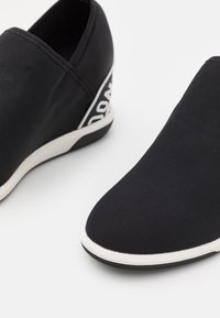 Baskets noires à enfiler avec semelles blanches et tige en mesh, comportant un texte blanc en guise de marque sur la partie talon.