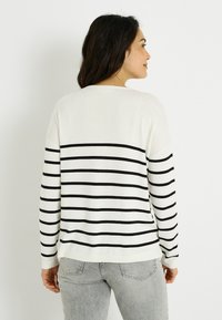 Jersey con rayas negras sobre fondo blanco, de corte holgado y mangas largas. El material parece suave, con un cuello redondo.