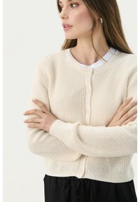 Beige neulecardigan, jossa on ribbetekstuuri, pyöreä kaula-aukko ja nappikiinnitys. Pitkät hihat ja rento malli. Valkoinen aluspaita näkyvissä.