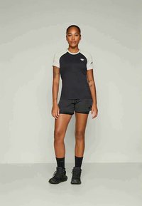 Γυναίκα στέκεται μετωπικά φορώντας μαύρο και άσπρο αθλητικό πουκάμισο κοντό μανίκι, μαύρη σορτς, μαύρες κάλτσες και μαύρα αθλητικά παπούτσια σε απλή φόντο.