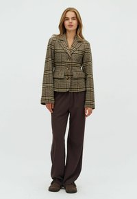 Blazer a quadri marrone con vestibilità strutturata, ampi rever e tre bottoni dorati, abbinato a pantaloni marroni a gamba larga e scarpe marroni.