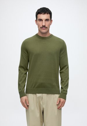 Mand med kort mørkt hår og overskæg iført en olivengrøn langærmet sweater og lyse beige vidde bukser, stående mod en ensfarvet baggrund.