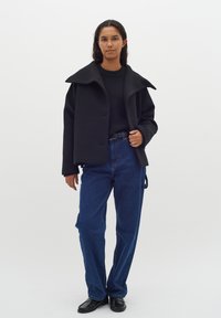 Cappotto di lana nero con un colletto oversize, indossato sopra un maglione nero. Abbinato a jeans blu a vita alta e mocassini neri.